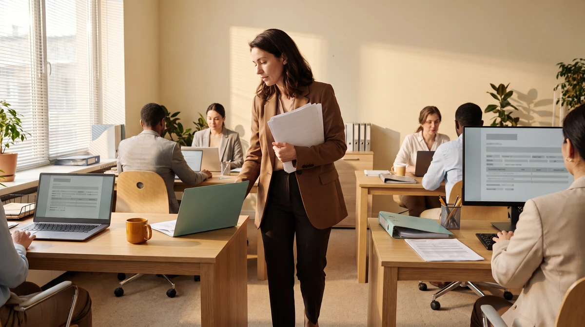 A coordinator crossing the floor with printed pages, stopping mid-sentence at a desk
